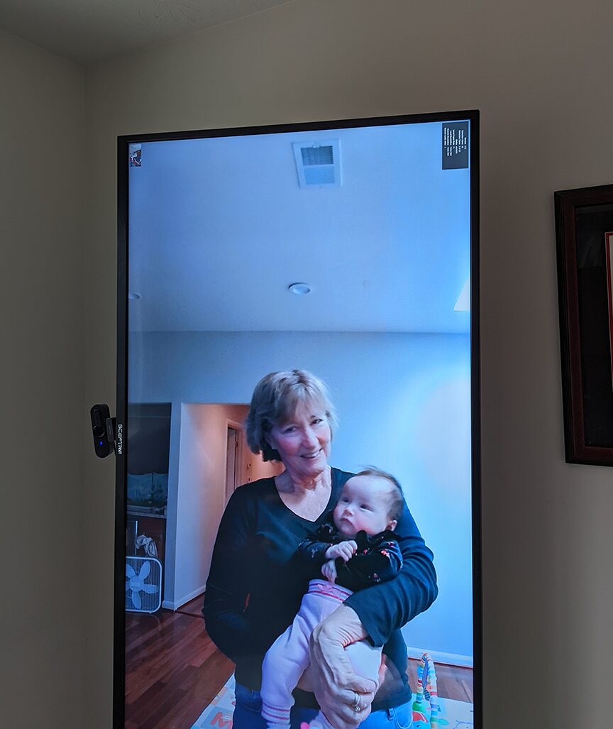 A portrait-mode TV on a wall showing a grandmother smiling and holding a baby in her living room.