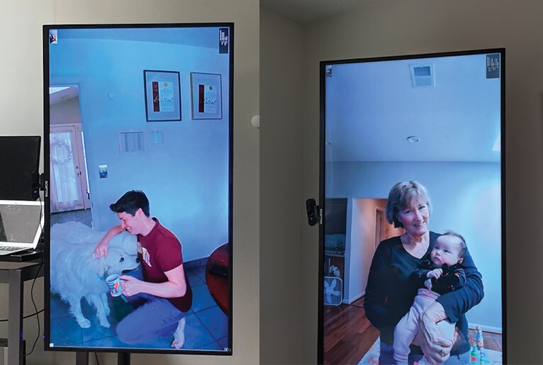 Two portrait-mode TVs mounted in two homes. One shows a man kneeling with a white dog, the other shows a grandmother holding a baby.